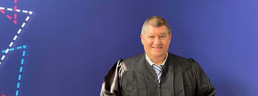 The image shows gradate William Murphy in black robes standing in front of a blue background. 
