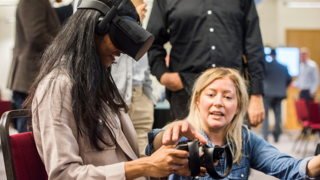 A female with long dark hair wearing a VR headset and holding a controller, assisted by a female with blonde hair