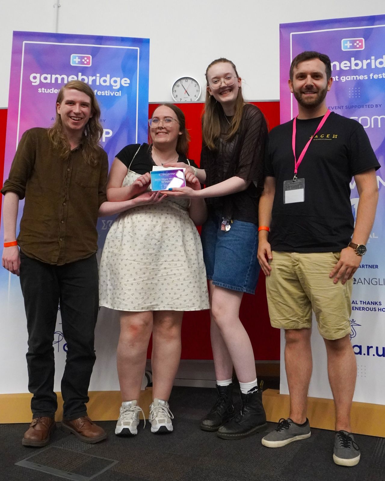 Three members of Riverboat Games accept their Best Team Project award on stage at Gamebridge. They are joined on stage by the award host. All of them smile to the camera.