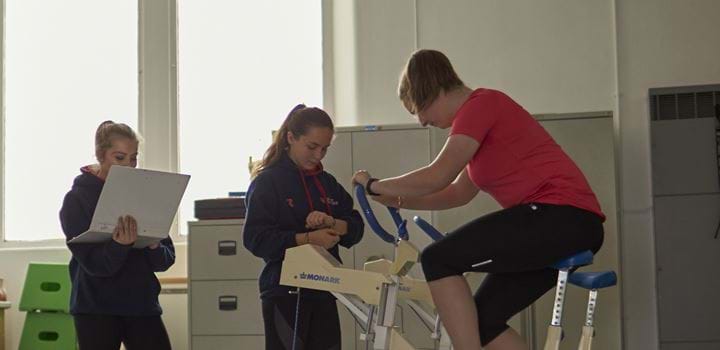 Female working on an exercise bike - two other females spectating and taking notes