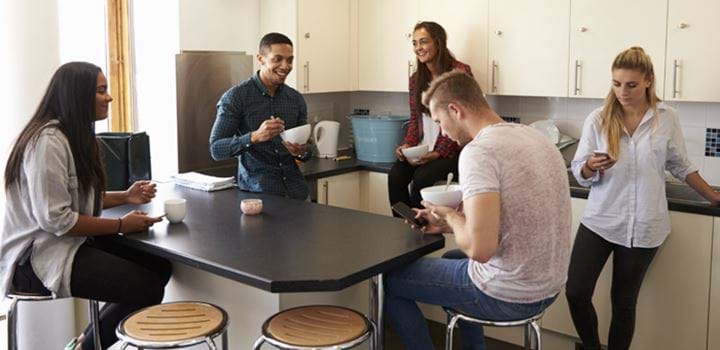 Group of five young adults huddled around a breakfast bar in a kitchen, three girls and two guys, three of the young adults are looking at their smartphones.