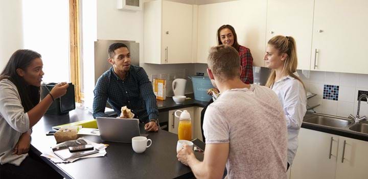 Group of five young adults huddled around in a kitchen, three girls and two guys. One of the guys is eating a sandwich, one of the girls is eating pasta, the other three students are not eating.