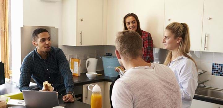 Group of four young adults huddled around in a kitchen, two girls and two guys. One of the guys is eating a sandwich.