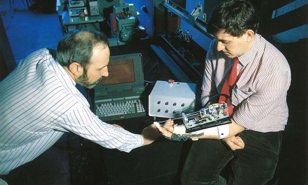 Photograph of two men sat in front of a computer at Dundee Institute of Technology (Abertay University). Man on the right has the hand therapy machine on his arm 