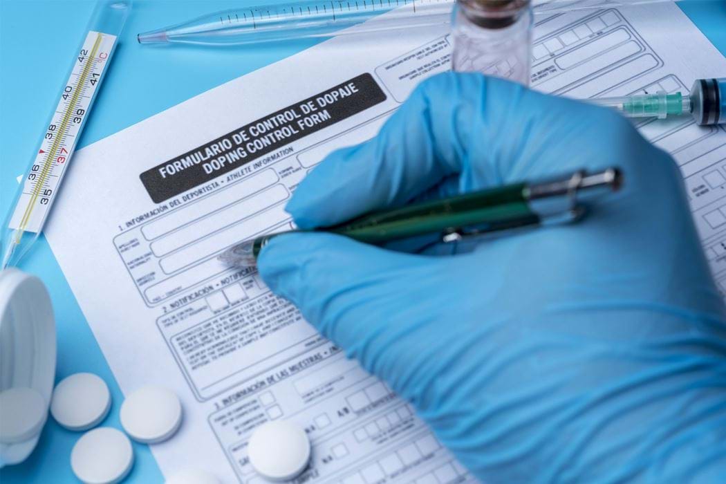 Plastic gloved hand writing doping control form, surrounded by drug paraphernalia