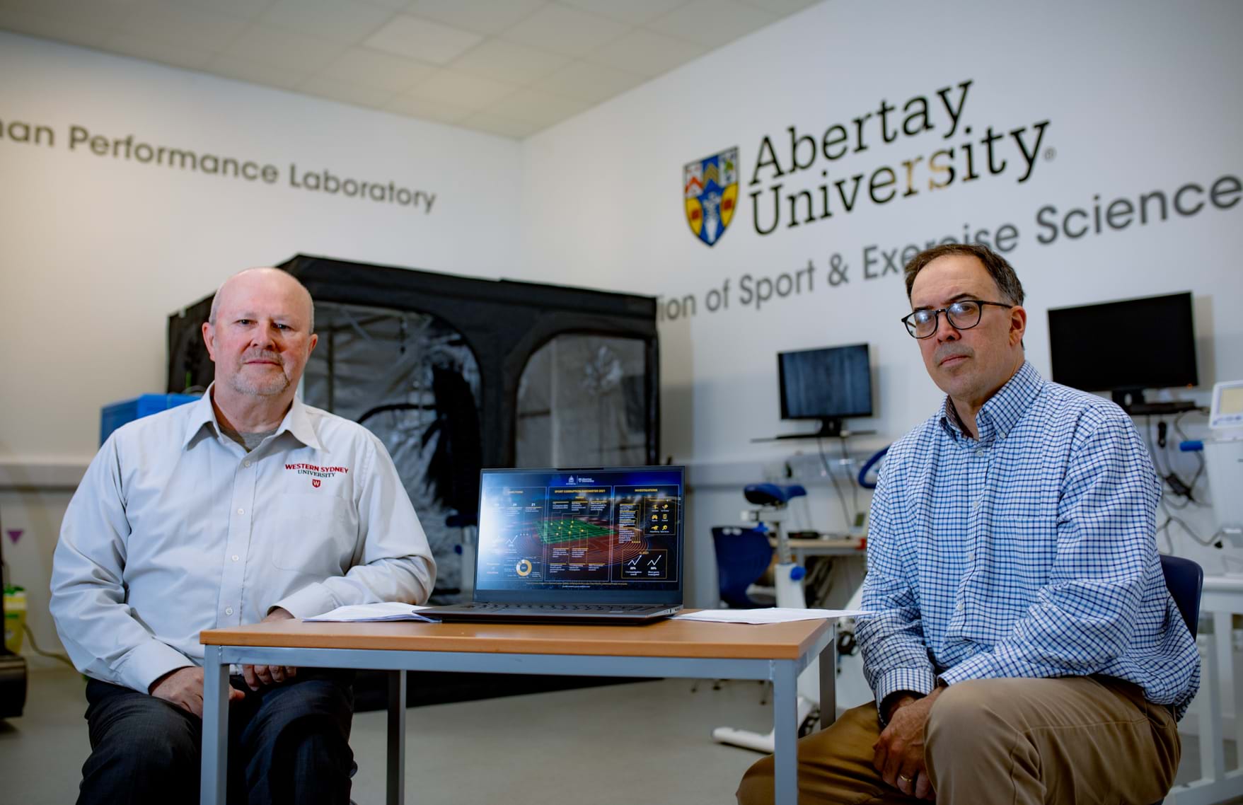 Neil Hall and David Lavallee sitting a sports lab with monitoring equipment in the background and Abertay University branding on the wall.