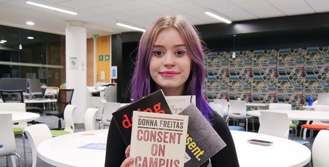 The image shows Student President Olivia Robertson in the Library holding up a range of books on Gender-Based Violence. - the book in the foreground is titled Consent on Campus.