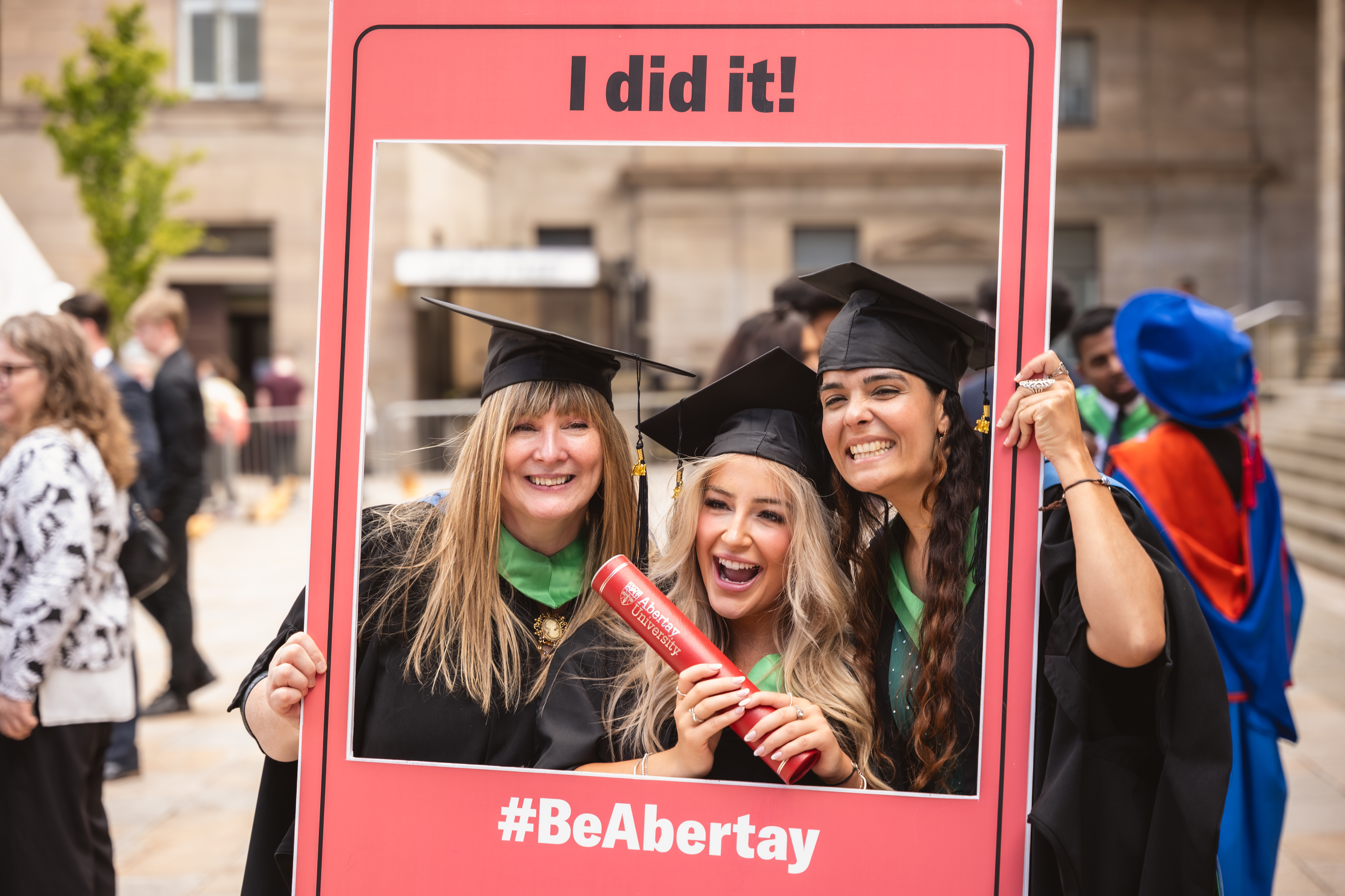 Three female graduates in gowns and hats pose with a large photo frame.