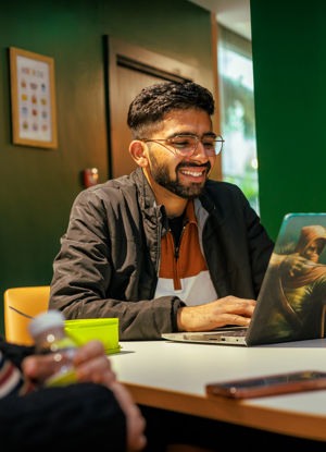 Male with short dark hair and glasses smiling while sitting in front of a laptop at a cafe table