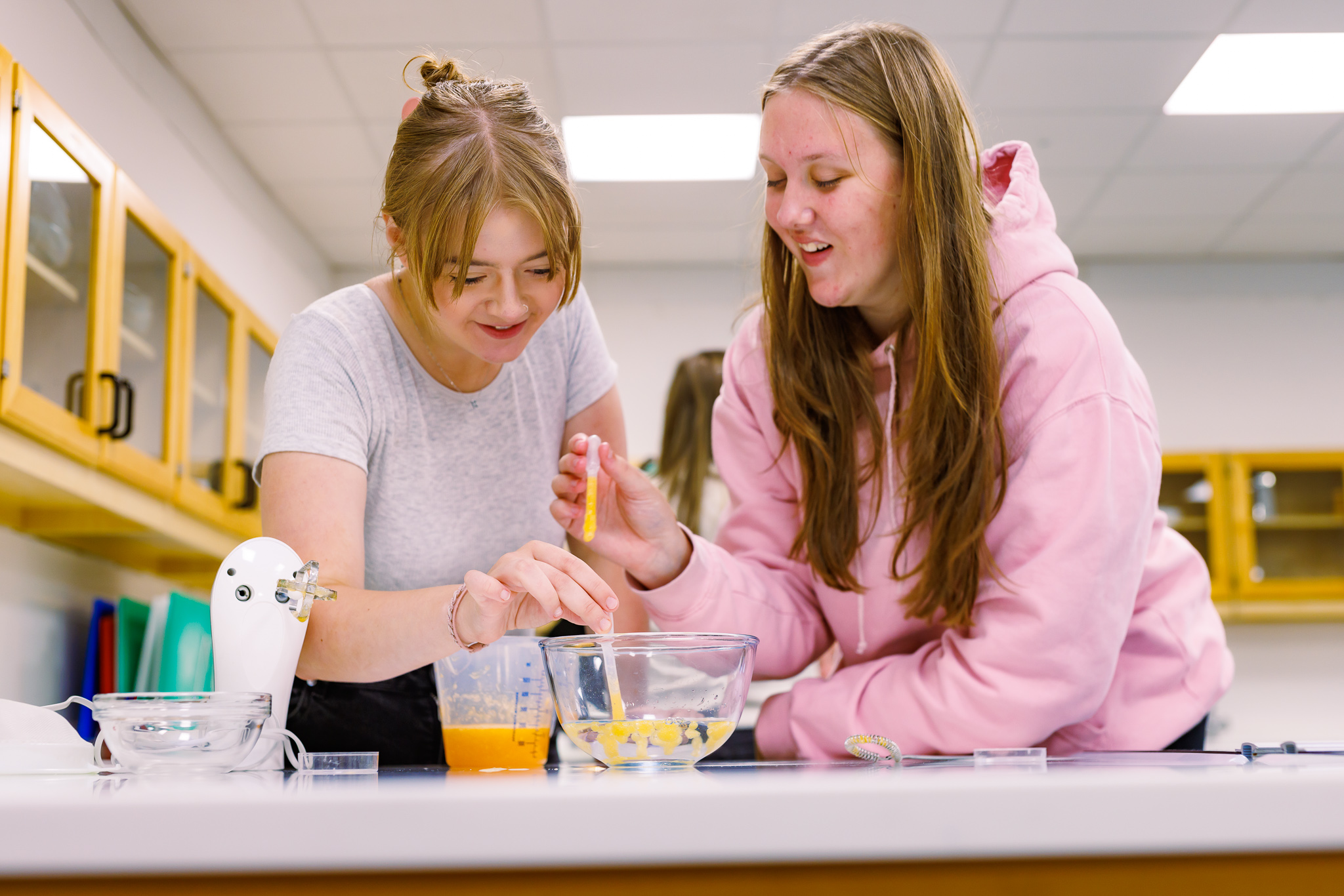 Two pupils from Kinross High School, one in a grey t-shirt and another in a pink hoody, smile as they create their own boba tea in one of the Abertay University food labs. They are stood over a clear food bowl producing the "bubbles" for the tea.