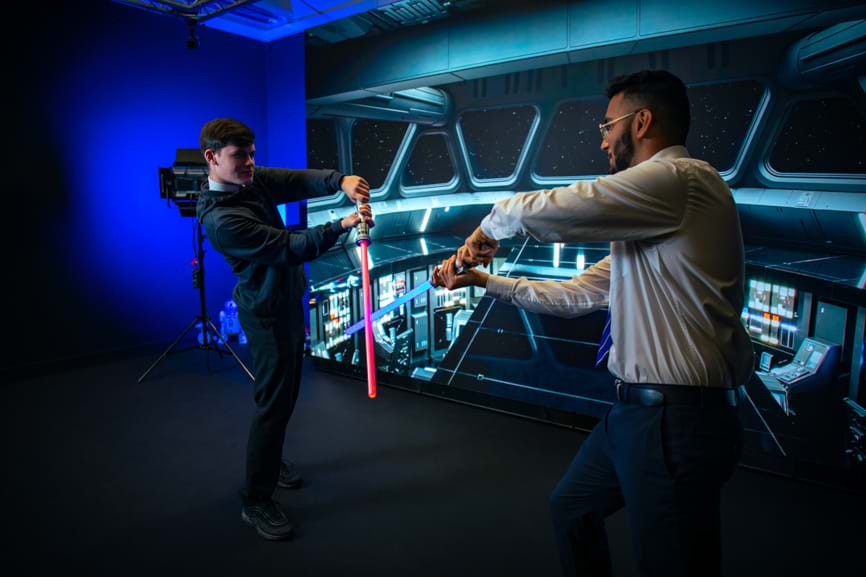 Two visiting school pupils play with toy light-sabers in Abertay University's Virtual Production Research Environment, in front of a screen displaying the inside of a Star Wars Star Destroyer ship.
