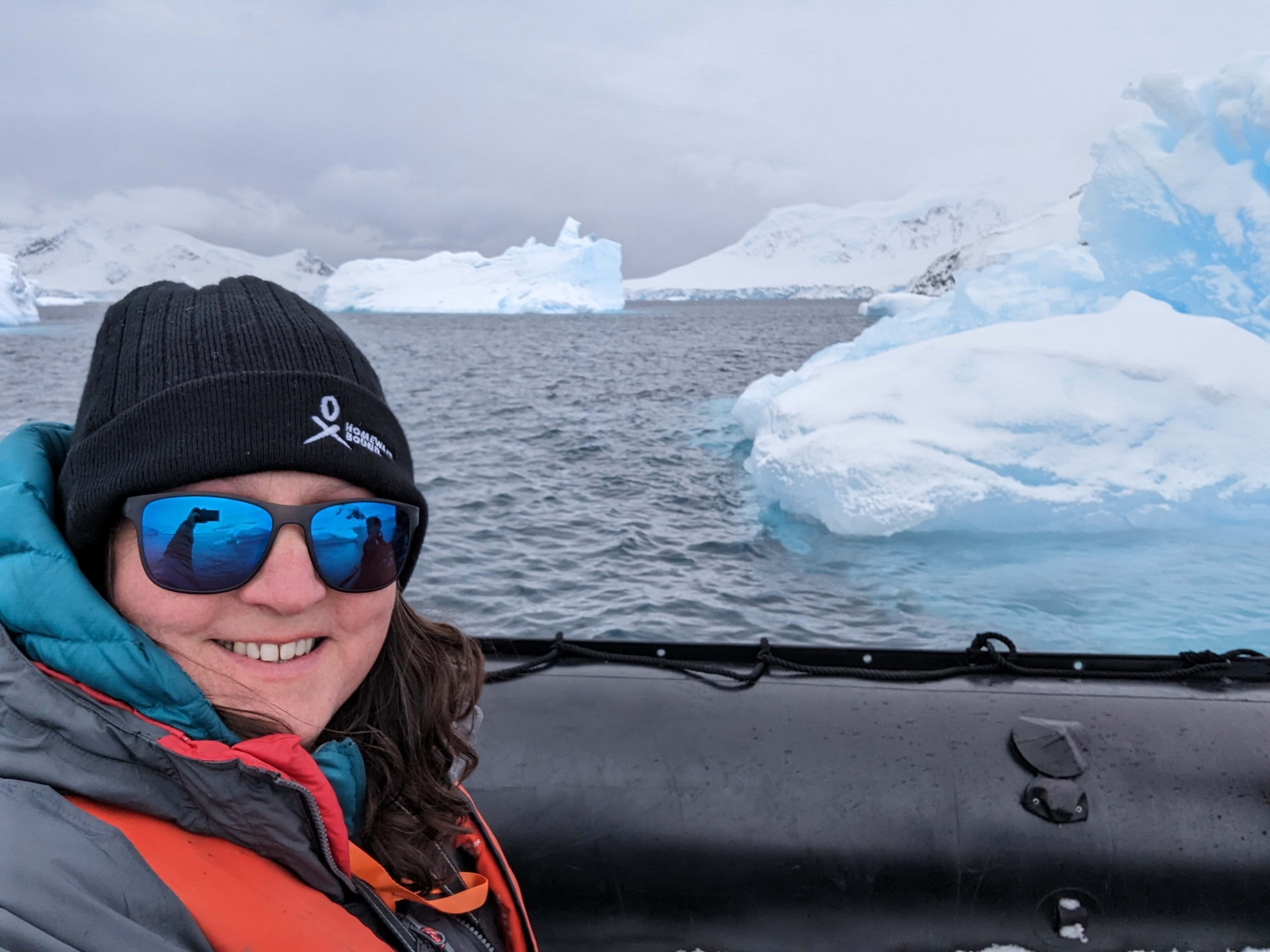 Abertay University lecturer Dr Rebecca Wade, wearing sunglasses and a hat, poses for s selfie on a boat in the Southern Ocean with icebergs floating in the sea behind her.